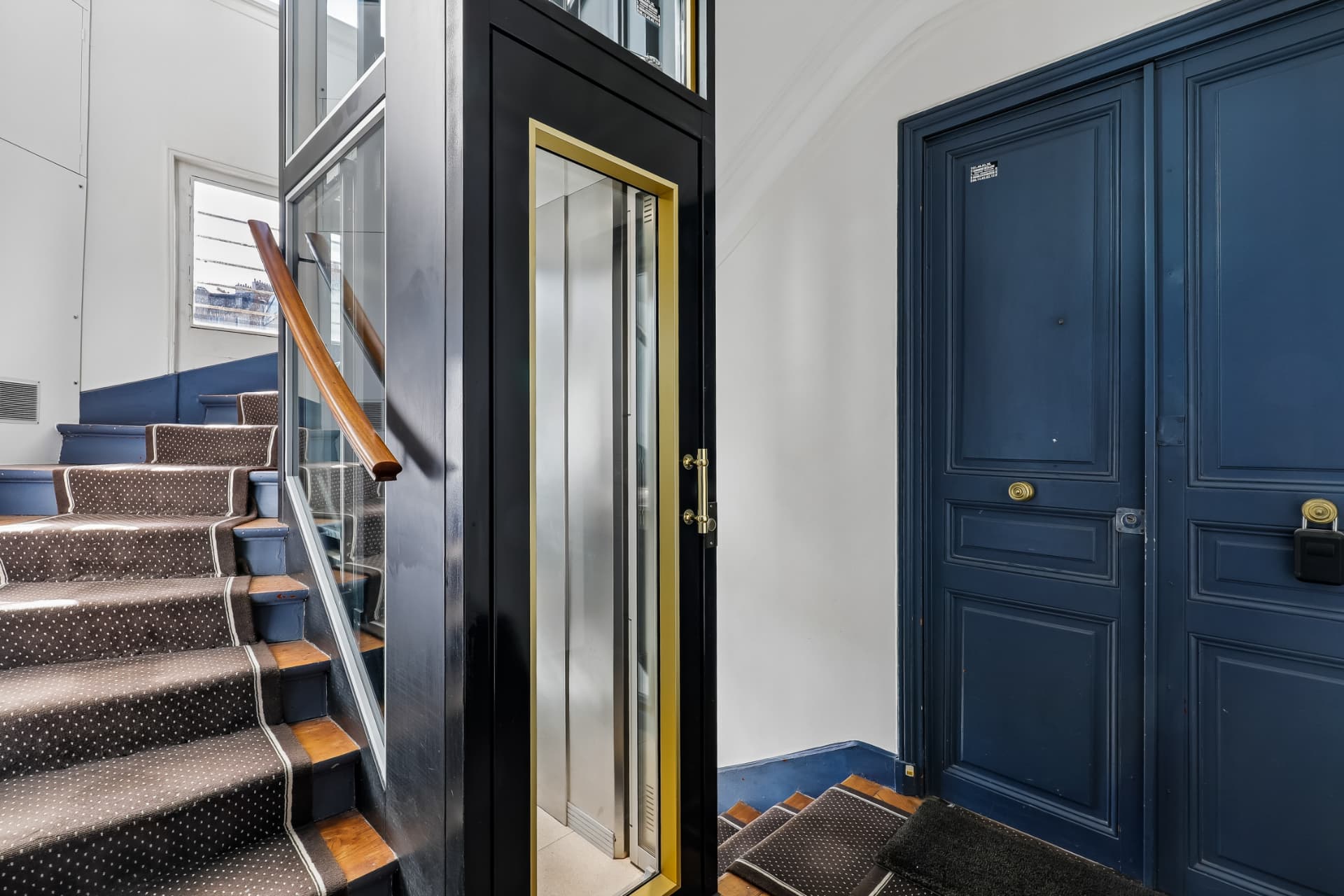 Cage d'escalier rénovée et ascenseur avec détails dorés, porte d'appartement bleu nuit